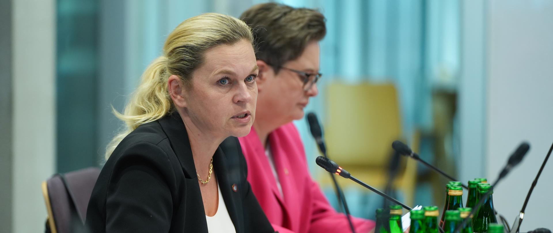 Minister Edukacji Barbara Nowacka przemawia przez mikrofon, siedząc przy stole. Przed nią stoją mikrofony, szklanki oraz butelki z wodą. Obok niej siedzi sekretarz stanu w MEN Katarzyna Lubnauer.