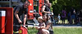 na zdjęciu strażak pompujący wodę hydronetka obok niego kobieta z dzieckiem, które celuje strumieniem wody w nalewak, w tle samochód gaśniczy przechodnie
