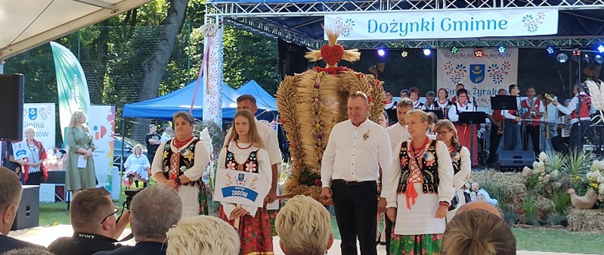 Grupa ludzi w strojach ludowych z tradycyjnym wieńcem dożynkowym. 