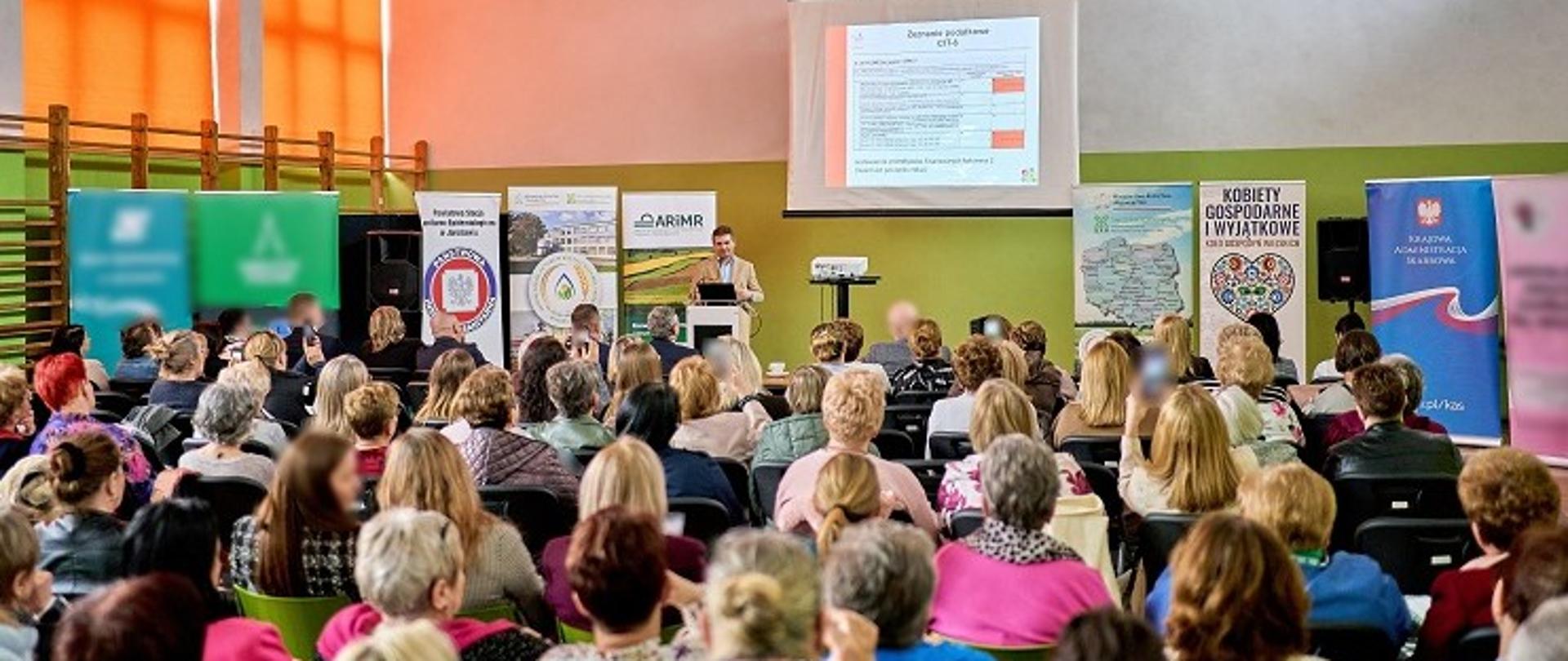 W sali siedzą licznie zgromadzeni uczestnicy szkolenia. Przed nimi stoi przy mównicy ekspert KAS.