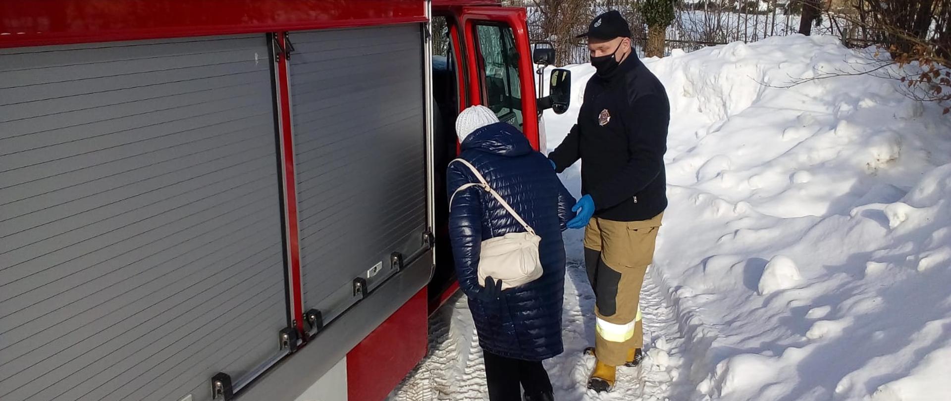 Zdjęcie przedstawia strażaka pomagającego kobiecie wejść do pojazdu pożarniczego, którym zostanie przetransportowana do punktu szczepień