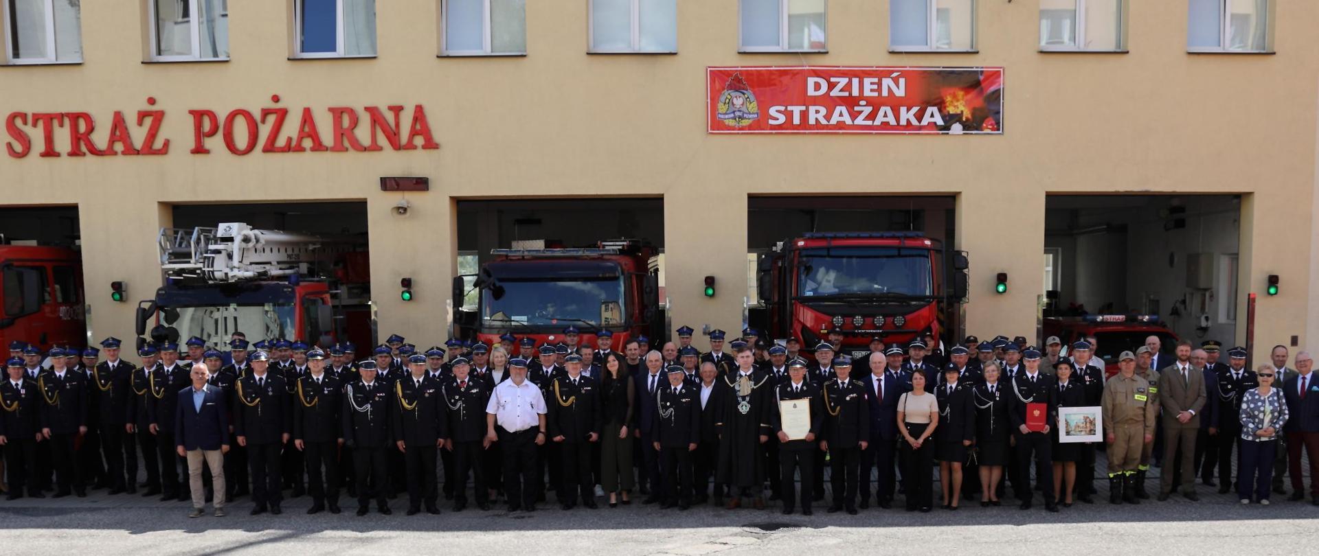 Zdjęcie przedstawia ustawionych przed budynkiem strażaków, na tle samochodów strażackich