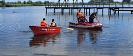 Na pierwszym planie łódź ratunkowa OSP Jutrosin oraz ponton OSP Pakosław na wodzie – jedna z załogą w czerwonych strojach, a druga z ratownikami w kamizelkach asekuracyjnych, którzy wiosłują. W oddali widać pomost oraz ratownika w wodzie.
