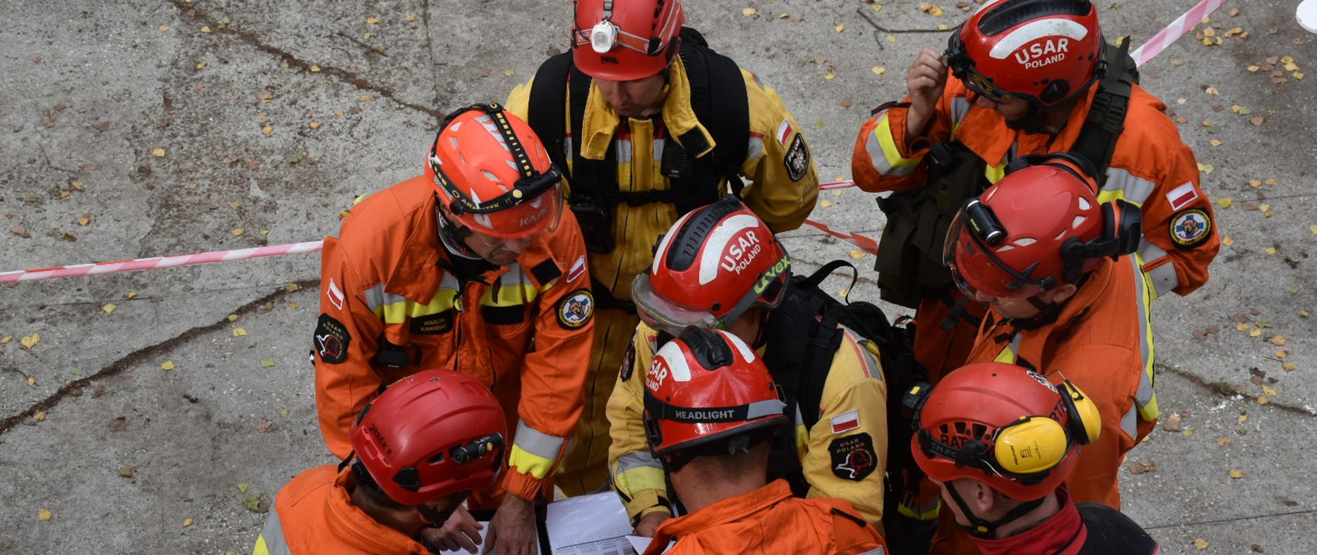 Strażacy w pomarańczowych kombinezonach, czerwonych kaskach stojący w kręgu spoglądają na kartkę zawierającą informację o planowanych ćwiczeniach