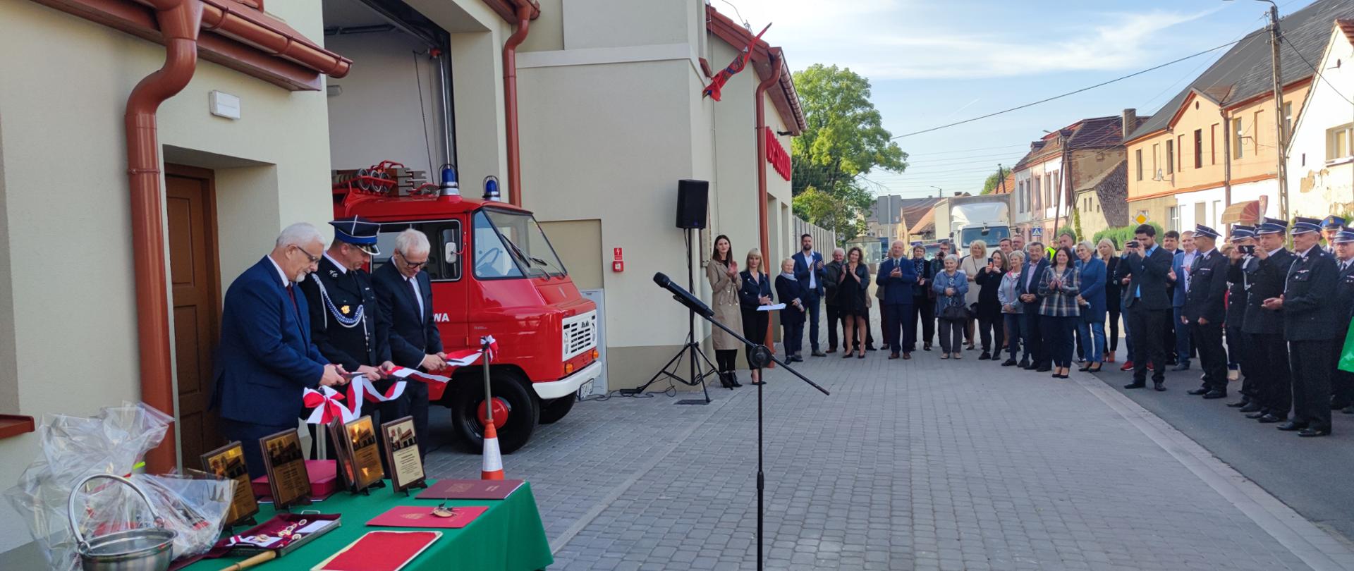 na zdjęciu widać remizę ochotniczej straży pożarnej w Sobocisku. Po lewej stronie są przecinający wstęgę: Henryk Kuriata, druh Łukasz Ostropolski i wojewoda dolnośląski Jarosław Obremski. Po prawej stronie widać licznie zgromadzonych gości.
