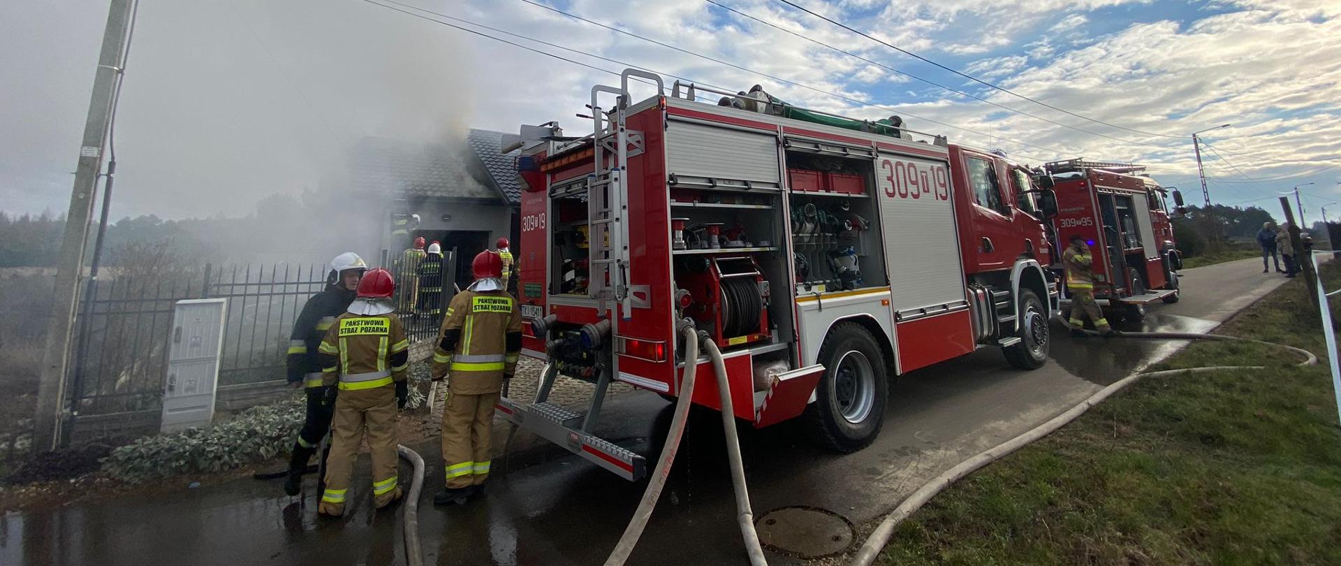 Na pierwszym planie widać dwa samochody pożarnicze z podpiętymi wężami gaśniczymi. Przy wozach stoją strażacy. Głębiej dom jednorodzinny gdzie paliła się więźba dachowa nad garażem – tam pracują strażacy podając wodę na ognisko pożaru.