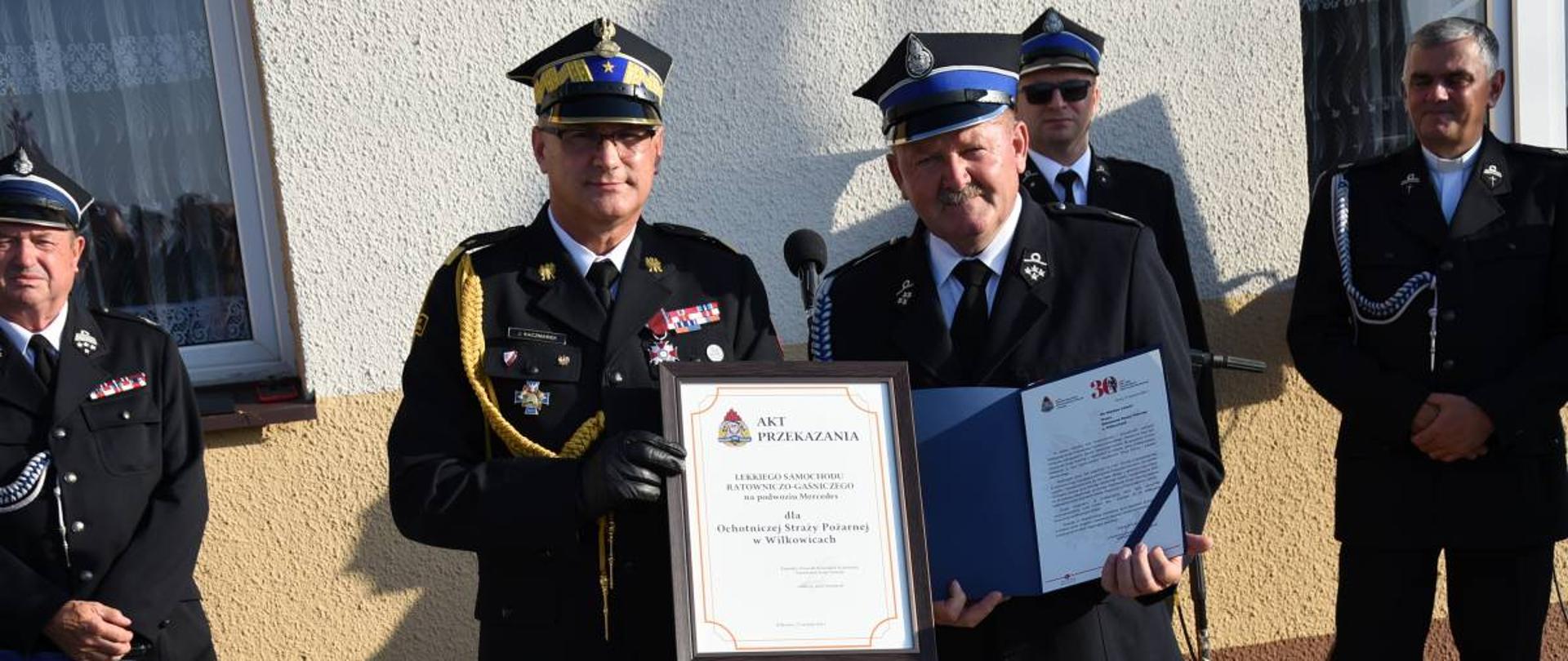 Komendant wojewódzki i prezes OSP Wilkowice pozują z prezentowanymi w dłoniach dokumentami. Obaj w mundurach wyjściowych. W tle goście uroczystości i budynek remizy.
