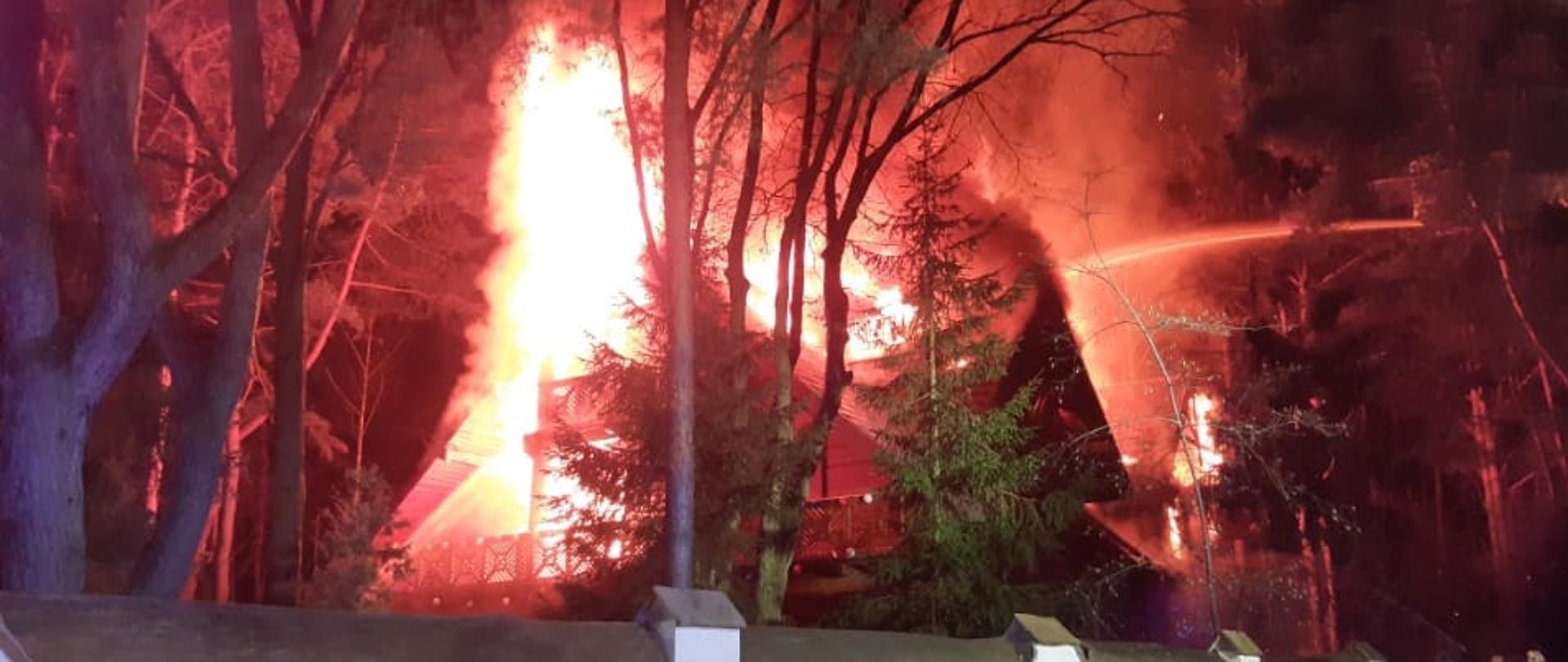 Noc. Drewniany budynek pali się, płomienie obejmują cały dach i poddasze. Widać rozwinięte węże oraz kosz podnośnika hydraulicznego, z którego podawany jest jeden prąd wody na palący się budynek. Dom stoi na posesji zadrzewionej. Drzewa od płomieni zabarwione są na czerwono i pomarańczowo.