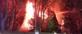 Noc. Drewniany budynek pali się, płomienie obejmują cały dach i poddasze. Widać rozwinięte węże oraz kosz podnośnika hydraulicznego, z którego podawany jest jeden prąd wody na palący się budynek. Dom stoi na posesji zadrzewionej. Drzewa od płomieni zabarwione są na czerwono i pomarańczowo.