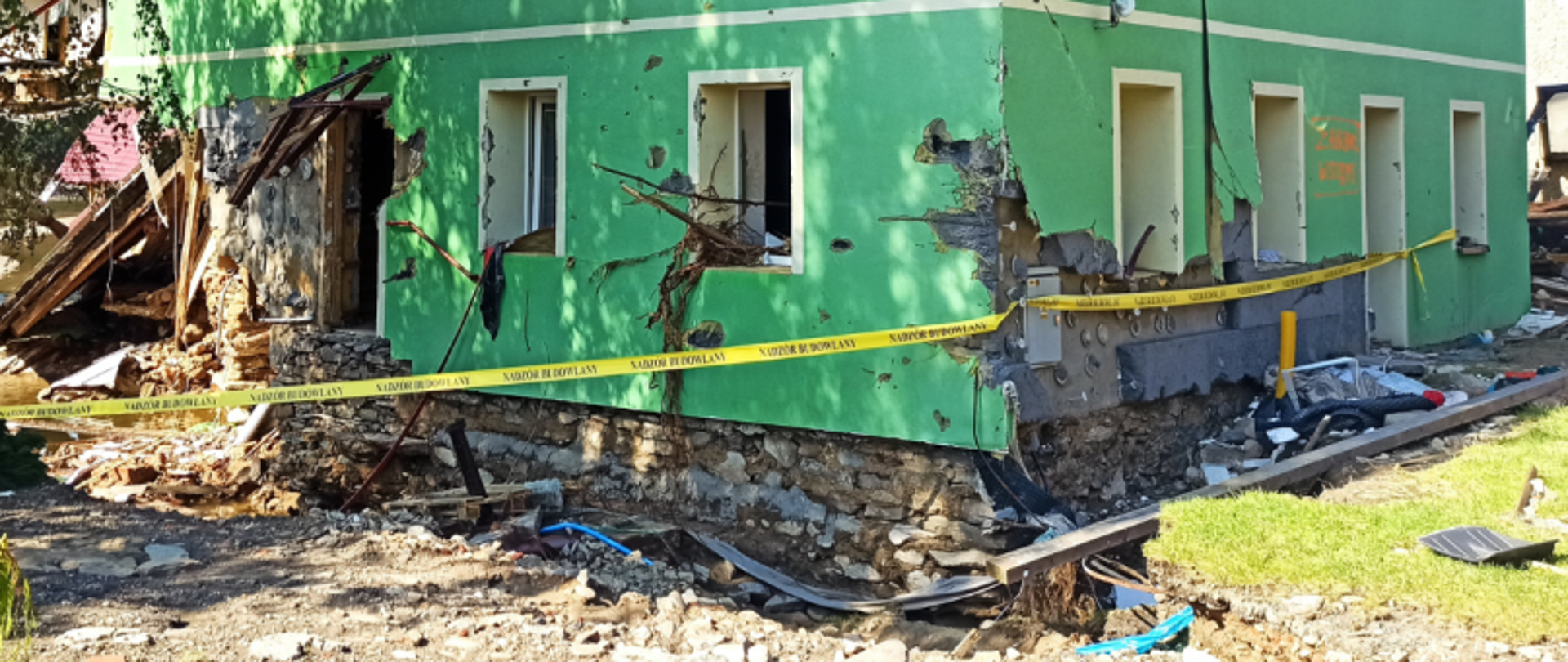 Zielony budynek z zawaloną elewacją, pęknięciami ścian i gruzem, otoczony taśmą ostrzegawczą.