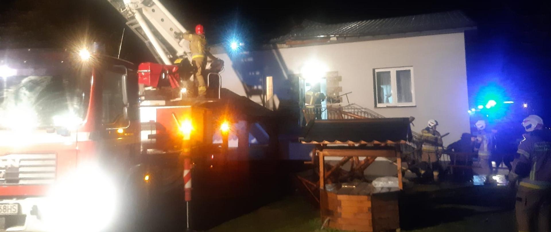 Na zdjęciu widać budynek, z którego zerwany częściowo jest dach. Przed budynkiem stoi strażacki podnośnik, który jest rozkładany. Ma włączone światła drogowe oraz żółte ostrzegawcze. Z boku budynku stoi inny samochód z włączonymi niebieskimi światłami na dachu. Na podwórku, przed budynkiem, widać strażaków w żółtych strojach oraz białych i czerwonych hełmach. Na środku podwórka jest studnia. Jest ciemno.