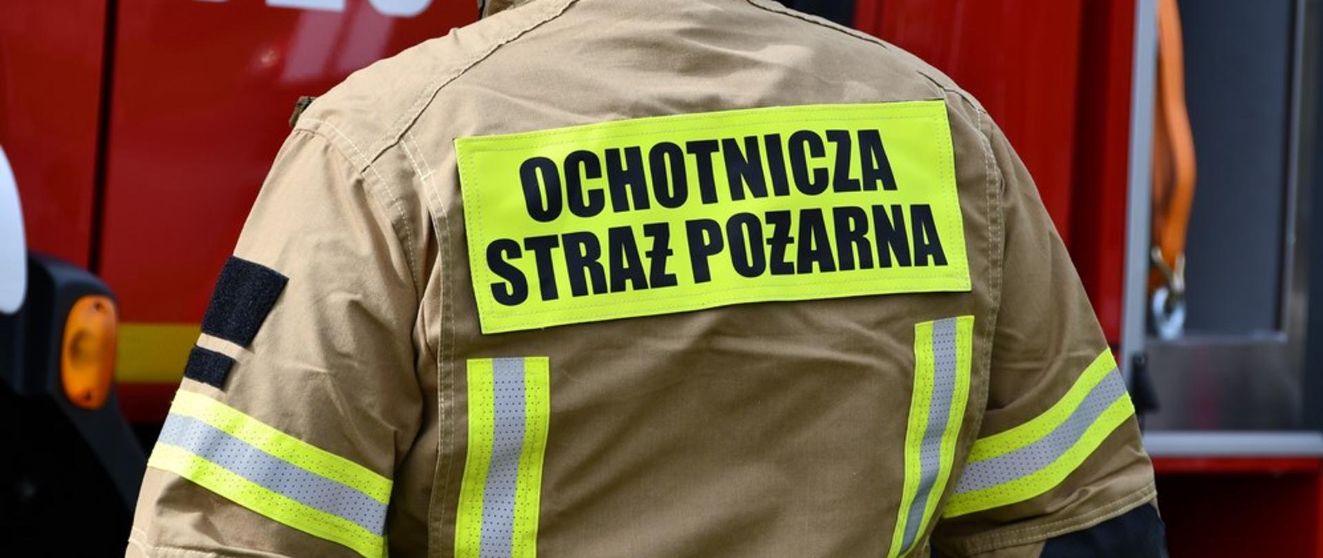 Zdjęcie przedstawia strażaka ochotniczej straży pożarnej stojącego tyłem od obiektywu na tle czerwonego samochodu pożarniczego. Na plecach strażaka widoczny napis ochotnicza straż pożarna.