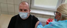 Zdjęcie przedstawia funkcjonariusza PSP, który znajduje się w gabinecie lekarskim i otrzymuje szczepionkę przeciwko COVID-19. Na zdjęciu jest również pielęgniarka ubrana w niebieskie ubranie ochronne oraz rękawiczki, która szczepi funkcjonariusza. Mężczyzna ubrany jest w ubranie dowódczo-sztabowe PSP. Obie osoby mają na sobie maseczki chirurgiczne.