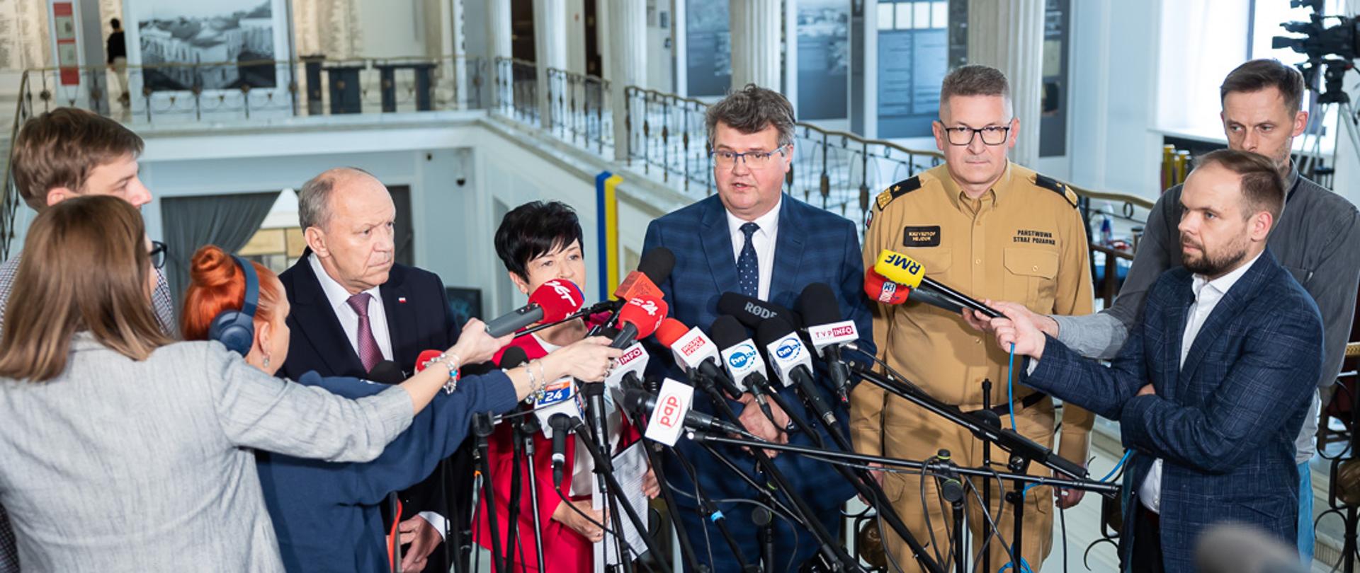 Wiceminister Maciej Wąsik podczas swojej wypowiedzi w sejmie RP przed mediami. Przed Wiceministrem stoją dziennikarze oraz rozstawione są mikrofony stacji radiowych i telewizyjnych. Obok Wiceministra z prawej strony stoi zastępca komendanta głównego PSP nadbryg. Krzysztof Hejduk, a z lewej przewodniczący Parlamentarnego Zespołu Strażaków Zbigniew Chmielowiec oraz poseł Joanna Borowiak.
