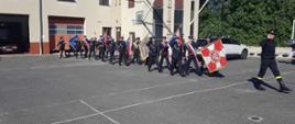 Szkolenie pocztów sztandarowych. Zdjęcie przedstawia plac komendy w Rawiczu. Na placu maszerują jeden za drugim, poczty sztandarowe OSP. Na ich czele strażak-dowódca z PSP. W tle budynek komendy.
