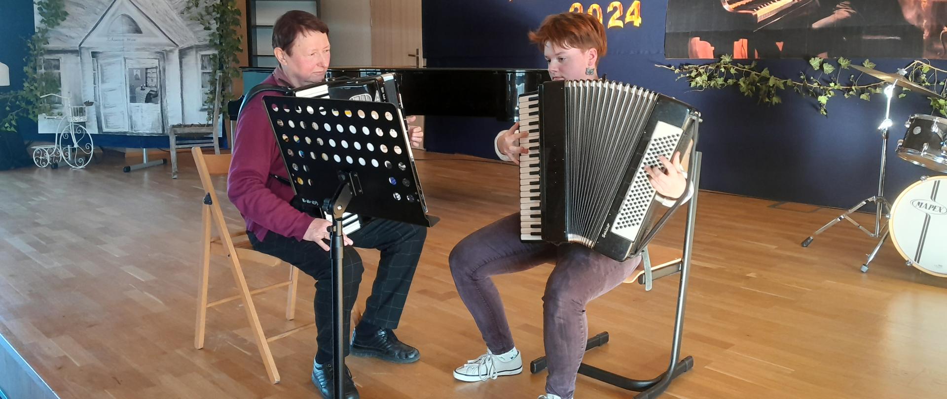 Na sali koncertowej z akordeonami siedzi przy pulpicie prof. Elżbieta Rosińska oraz uczennica Zofia Dzienisz. W tle widać dekorację pozostałą po niedawno obchodzonym Dniu Patrona