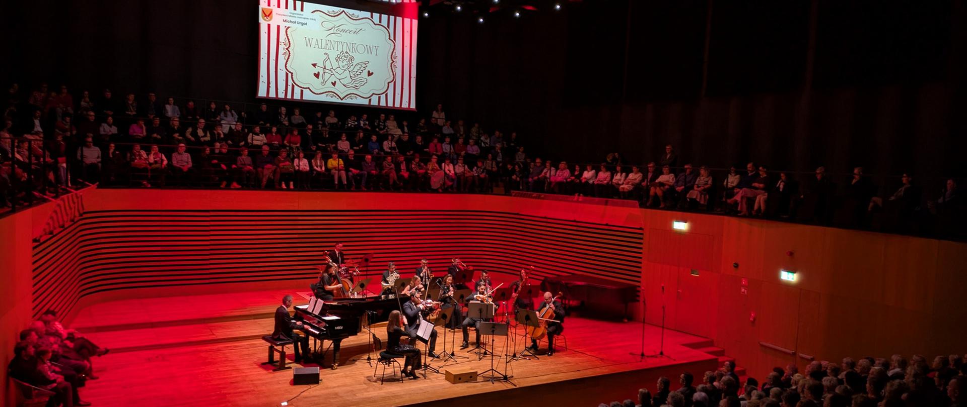 Na scenie gra orkiestra kameralna, mężczyzna gra na fortepianie. Z przodu i nad sceną widać publiczność.