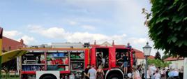 Na zdjęciu widzimy samochód strażacki oraz strażak pokazuje wyposażenie wozu. W tle chodzą ludzie i bawią się dzieci. 