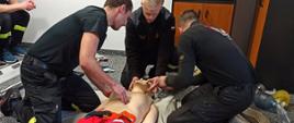 Zdjęcie w sali szkoleniowej. Trzech strażaków ćwiczy udzielanie pierwszej pomocy. Strażacy ćwiczą na fantomie. Dookoła widać rozłożone przedmioty służące do udzielania pierwszej pomocy