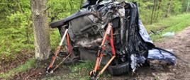 Na zdjęciu widoczny jest samochód leżący na lewym boku po dachowaniu. Samochód koloru niebieskiego, z widocznymi uszkodzeniami całej karoserii. Samochód od strony podwozia zabezpieczony podporami stabilizującymi w kolorze czerwonym. Z tyłu na tle samochodu widoczny las rozciągający się wzdłuż drogi.