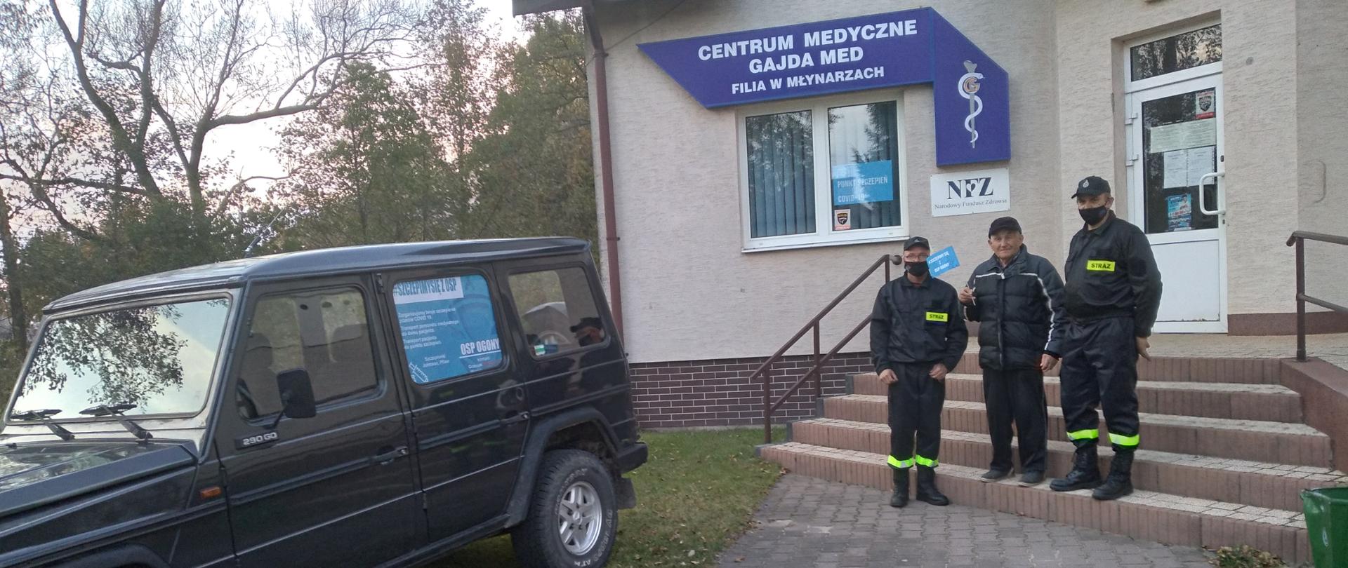 Dwóch strażaków OSP stoi przed przychodnią razem z mężczyzną w podeszłym wieku. Przed przychodnią stoi zaparkowany samochód OSP z logiem akcji #SzczepimySię z OSP