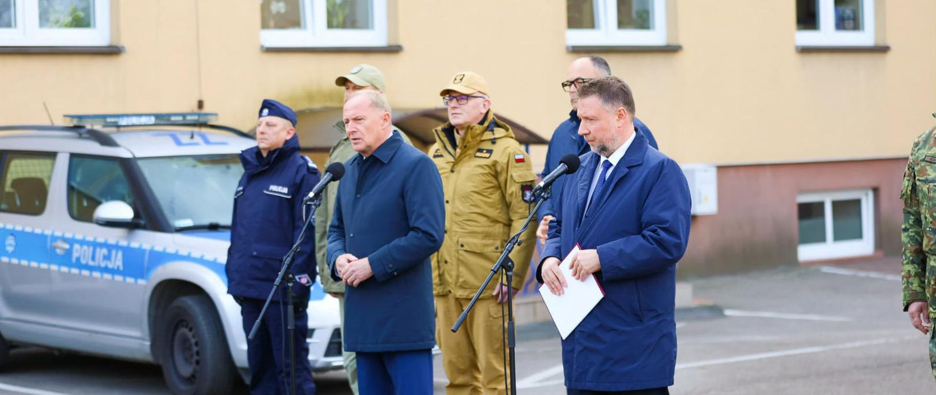 Minister Spraw Wewnętrznych i Administracji przemawia podczas briefingu prasowego w Komendzie Powiatowej Policji w Wołominie. Obok niego stoją funkcjonariusze Policji, Straży Granicznej oraz zastępca komendanta głównego PSP nadbryg. Paweł Frysztak. W tle widoczny radiowóz policyjny i budynek komendy.
