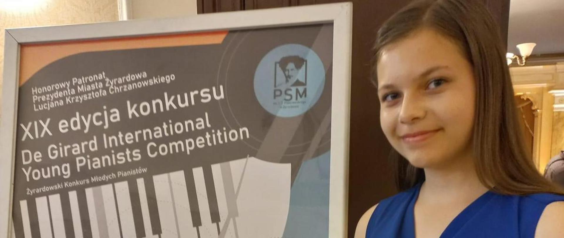 Dziewczyna w niebieskiej sukience stoi, w ręku trzyma dyplom, obok sztaluga z plakatem konkursu