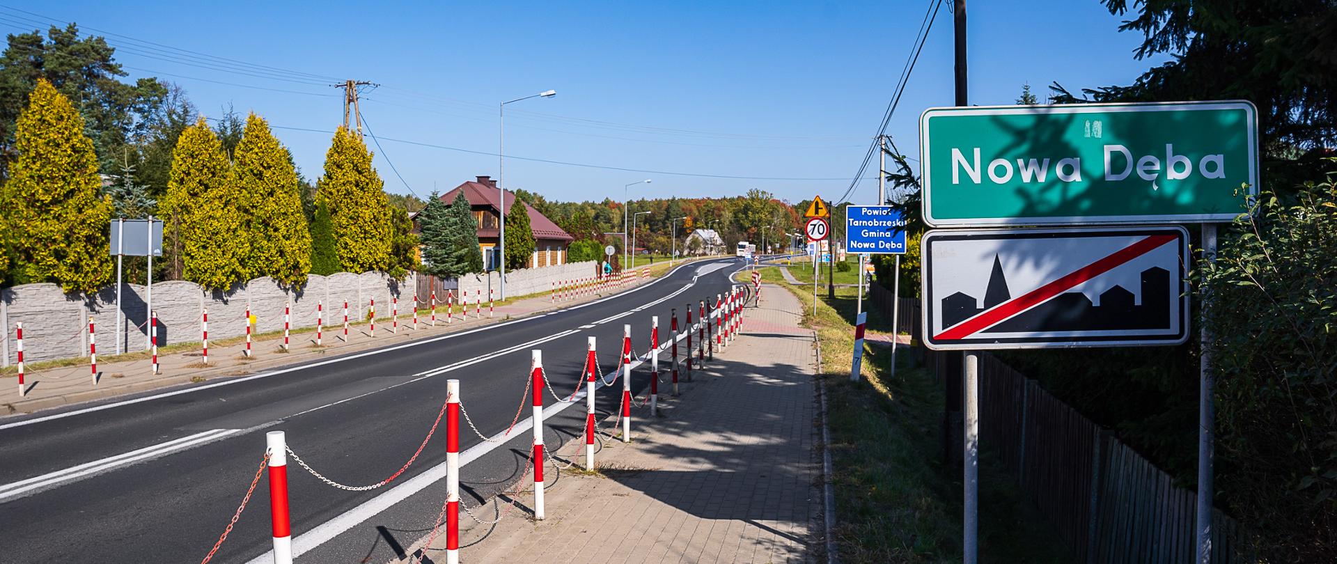 Na zdjęciu widoczny znak z napisem Nowa Dęba. Po lewej stronie widoczna jest droga i chodnik. Świeci słońce. 