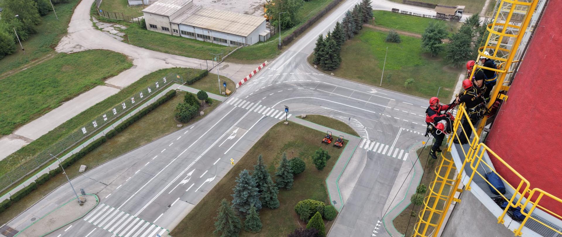 Fotografia przedstawia funkcjonariuszy podczas ćwiczeń: Strażacy stoją na galerii technologicznej, a jeden z ratowników zabezpiecza ewakuację osoby poszkodowanej w noszach. Daleko w dole stoją pojazdy strażackie.