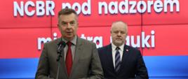 Na tle wbudowanego w ścianę wielkiego ekranu z napisem NCBR pod nadzorem Ministra Nauki stoi minister Wieczorek który mówi do mikrofonu na stojaku, obok niego wiceminister Gzik.