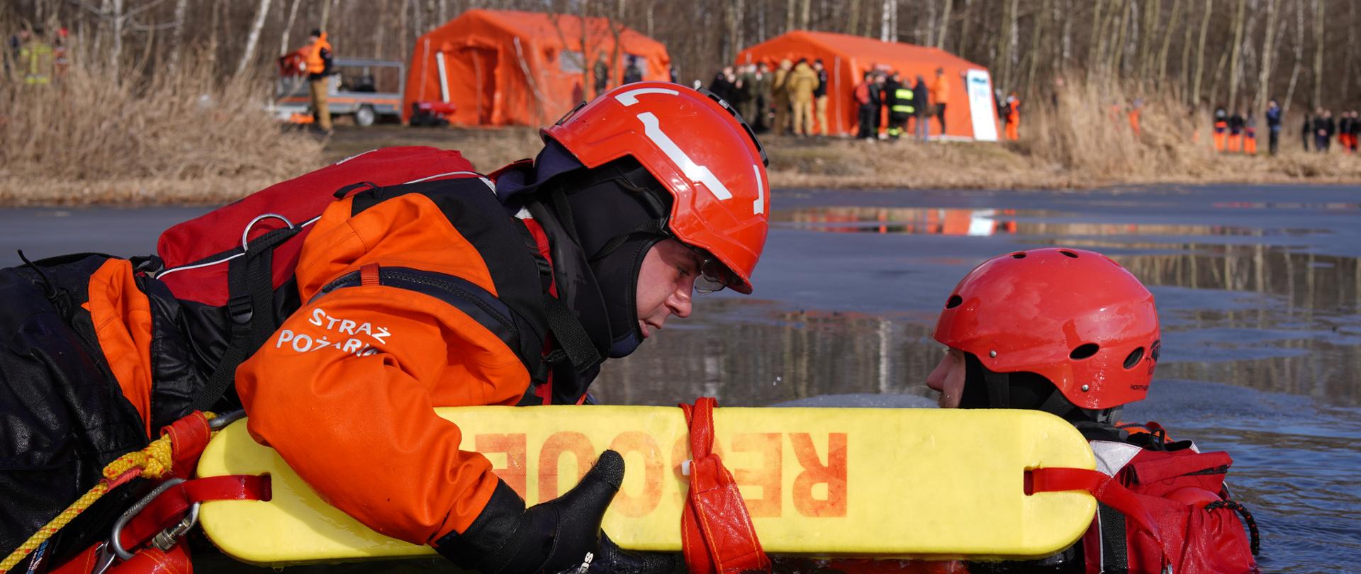 Dwóch ratowników w czerwonych kaskach i pomarańczowych kombinezonach w zimnej wodzie, prowadzą akcję ratunkową na zamarzniętym akwenie; w tle widać namioty.