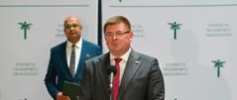 Wiceminister Rzymkowski stoi i mówi do mikrofonu, za nim stoi mężczyzna w niebieskim garniturze, w tle ścianka z napisem Inspekcja Transportu Drogowego.