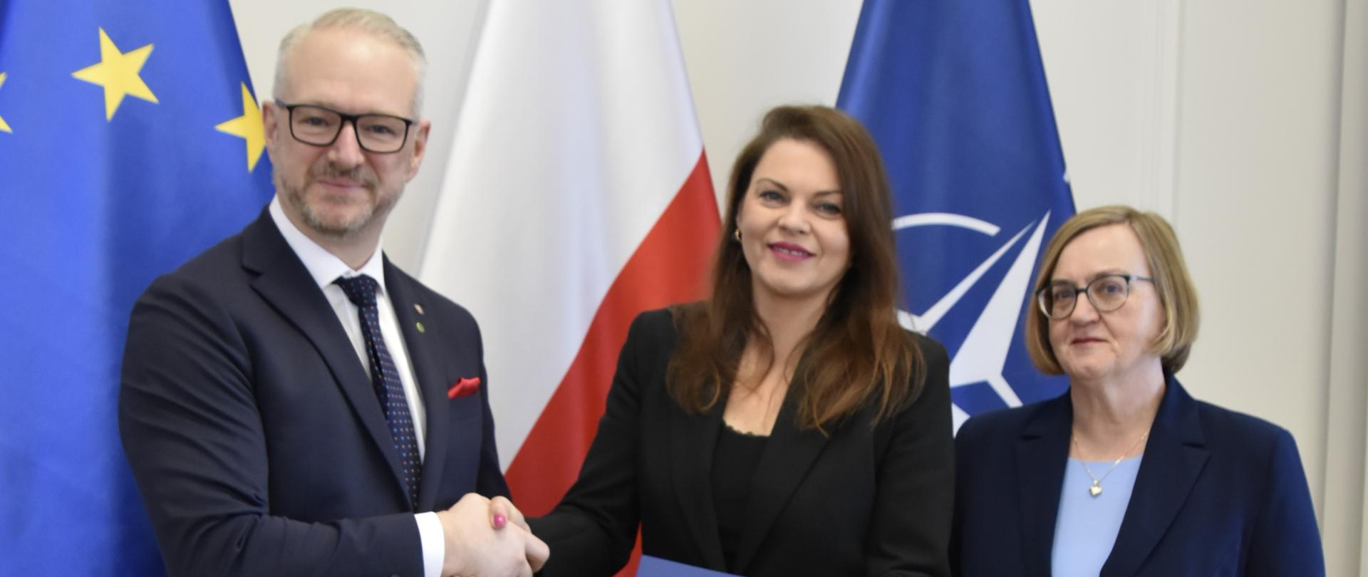 mężczyzna w garniturze podaje rękę kobiecie z ciemnymi włosami, usmiechają się 