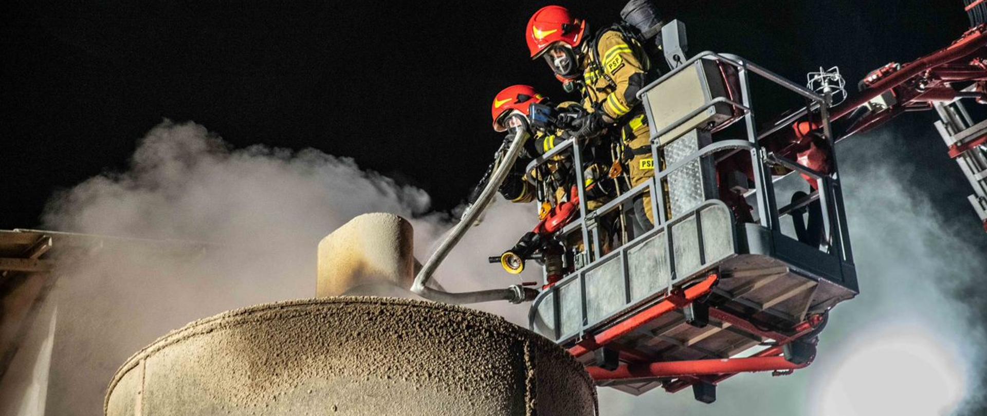 Zdjęcie przedstawia strażaków podczas gaszenia pożaru z góry suszarni do zboża. Strażacy znajdują się w koszu podnośnika hydraulicznego.
