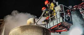 Zdjęcie przedstawia strażaków podczas gaszenia pożaru z góry suszarni do zboża. Strażacy znajdują się w koszu podnośnika hydraulicznego.