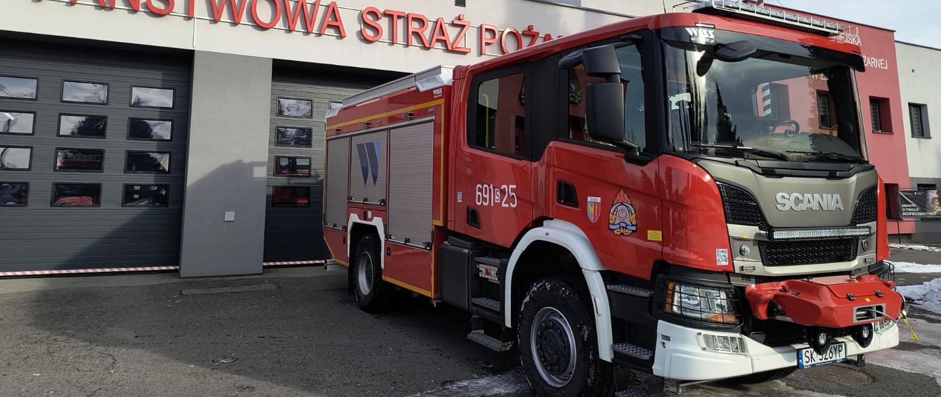 Samochód pożarniczy Scania ustawiony przed garażem komendy, nad nim napis Państwowa Straż Pożarna oraz powiewa flaga państwowa.