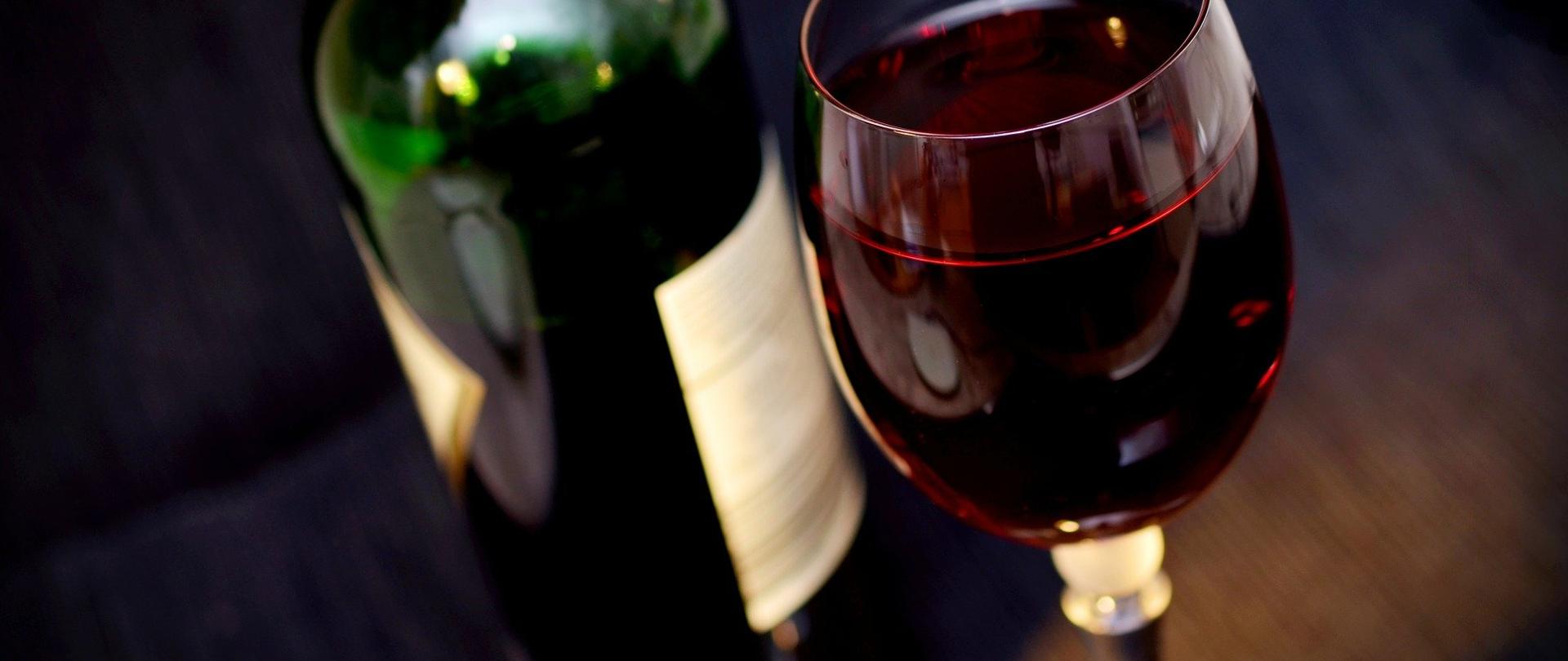 Na zdjęciu znajduje się butelka z czerwonym winem, obok której stoi kieliszek z czerwonym winem.