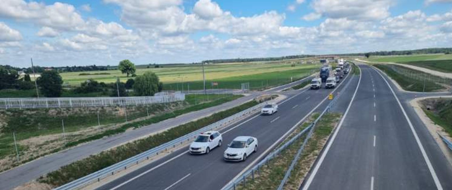 Nowa droga dwujezdniowa widziana z góry, z kilkoma samochodami jadącymi w jednym kierunku; w tle pola uprawne i błękitne niebo z rozproszonymi chmurami.
