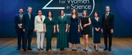 Na tle granatowej ściany z napisem 25 for Women in Science stoi trzech mężczyzn w garniturach i sześć kobiet w eleganckich sukniach.