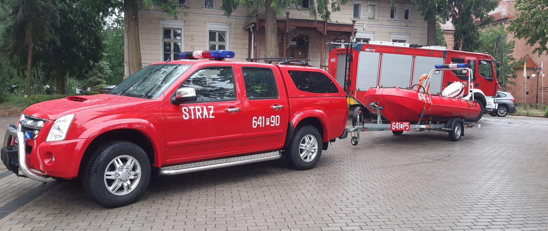 Na zdjęciu samochód strażacki z przyczepką, na której znajduje się łódź ratunkowa. W tle budynek ośrodka wypoczynkowego. Zdjęcie zrobione na zewnątrz.