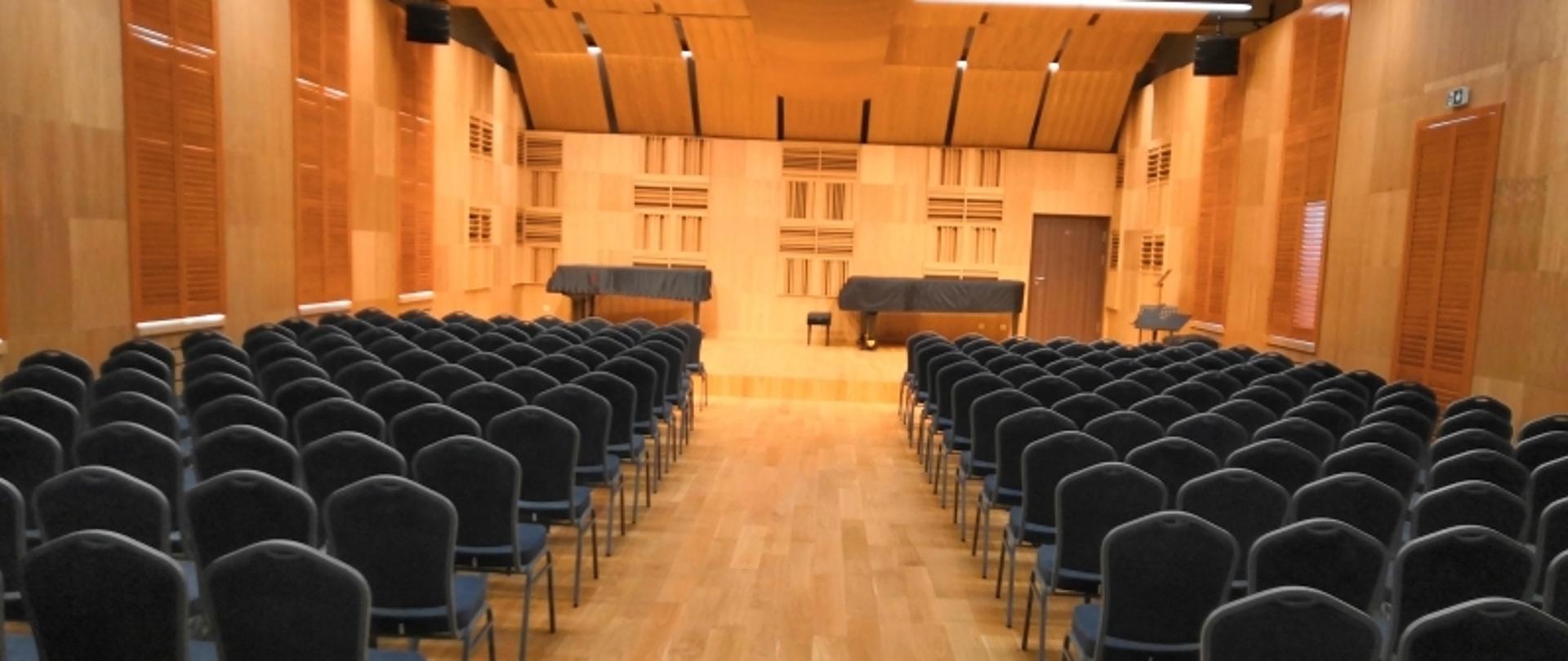 Sala koncertowa Zespołu Szkół Muzycznych w Pile. Widok od strony wejścia na estradę. Obszerna oświetlona sala, wykończona drewnem. Na suficie ekrany akustyczne. Rzędy krzeseł po obu stronach przejście środkiem do estrady na której stoją dwa fortepiany.