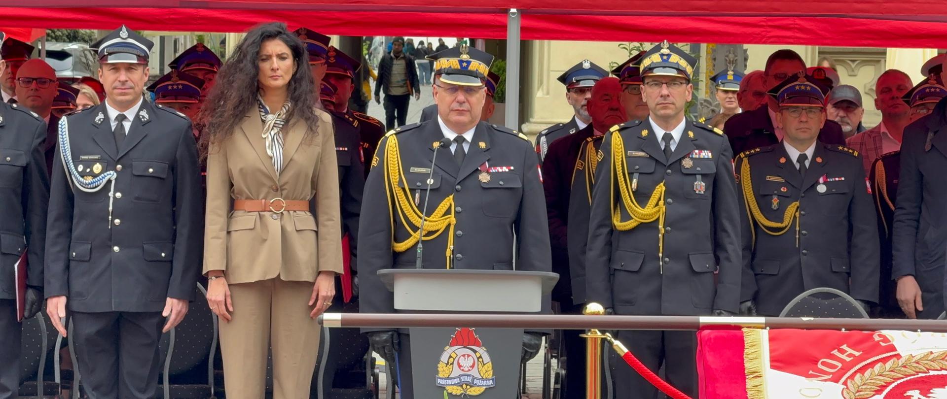 Przedstawiciele PSP i zaproszeni goście stoją pod czerwonym namiotem podczas oficjalnej części Wojewódzkich Obchodów Dnia Strażaka na wrocławskim Rynku.