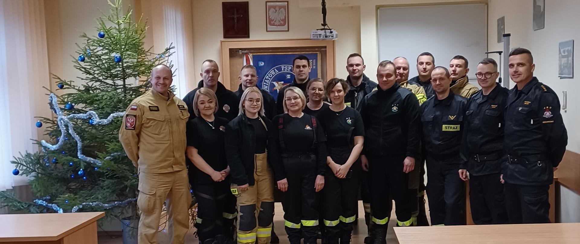 Druhny, druhowie i funkcjonariusze stoją na świetlicy obok siebie i wykonają pamiątkowe zdjęcie po zdanym egzaminie z KPP. Po lewej stronie widoczna jest ubrana choinka. 