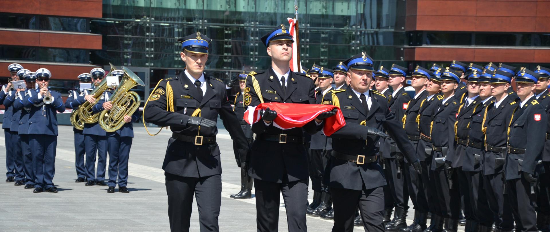 Trzech funkcjonariuszy PSP w galowych mundurach niesie złożoną biało-czerwoną flagę. Towarzyszy im orkiestra wojskowa grająca na instrumentach dętych. W tle szereg strażaków w umundurowaniu galowym.