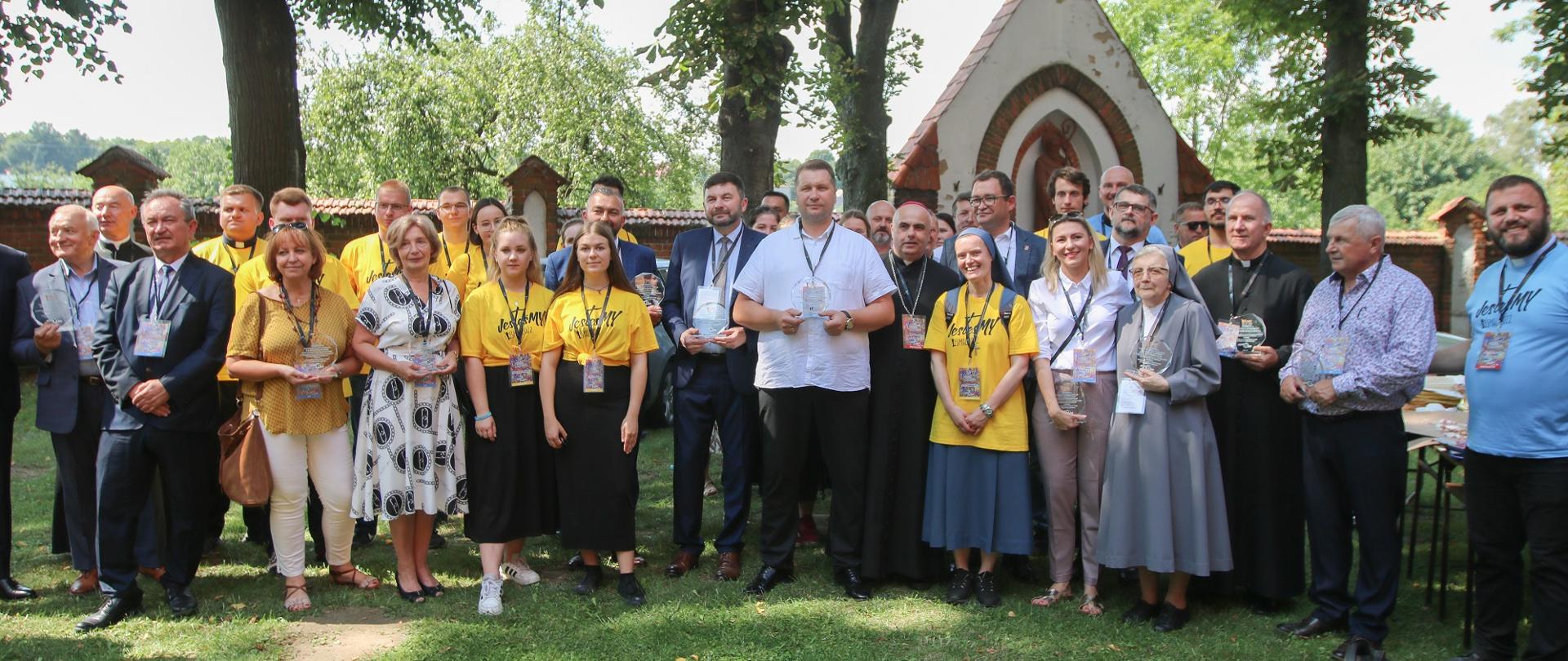 Zdjęcie zbiorowe - duża grupa ludzi stoi na trawie, za nimi kapliczka.