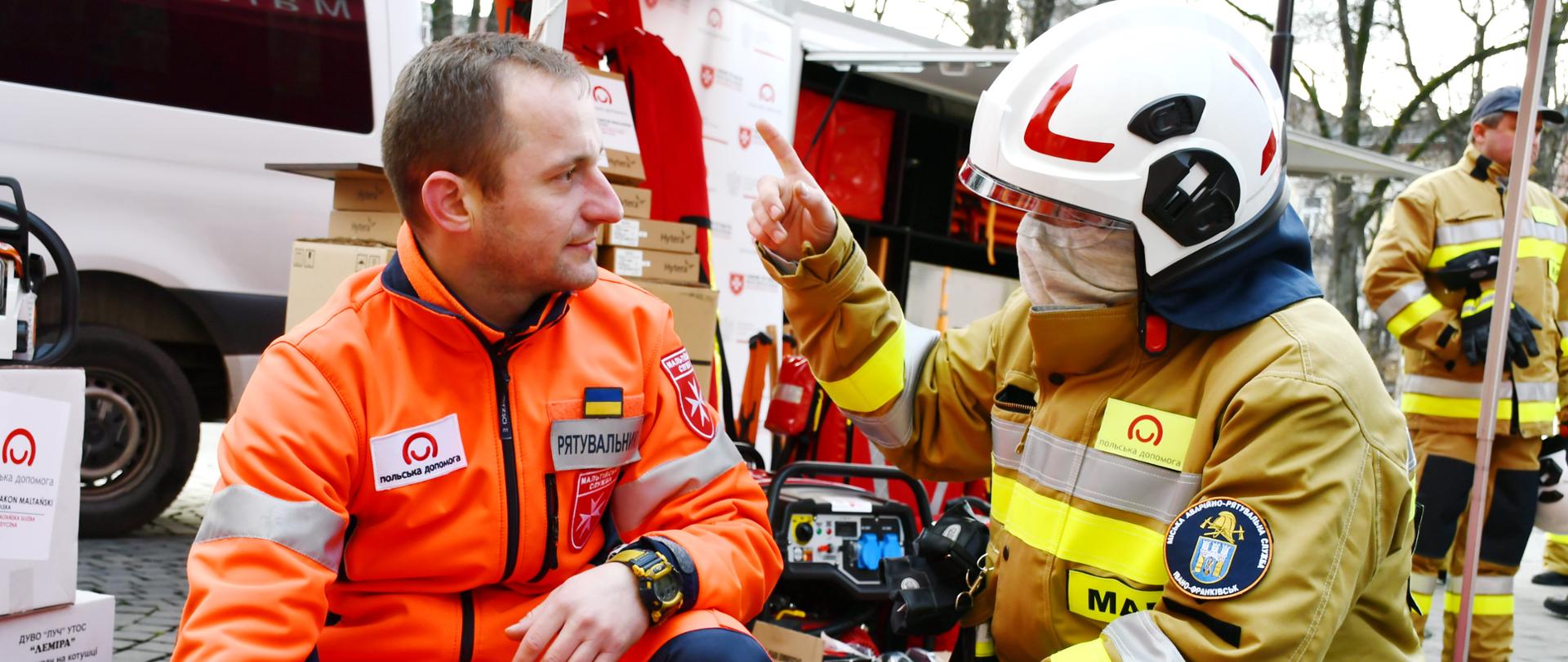 Strażak i ratownik medyczny kucają przy samochodzie strażackim.