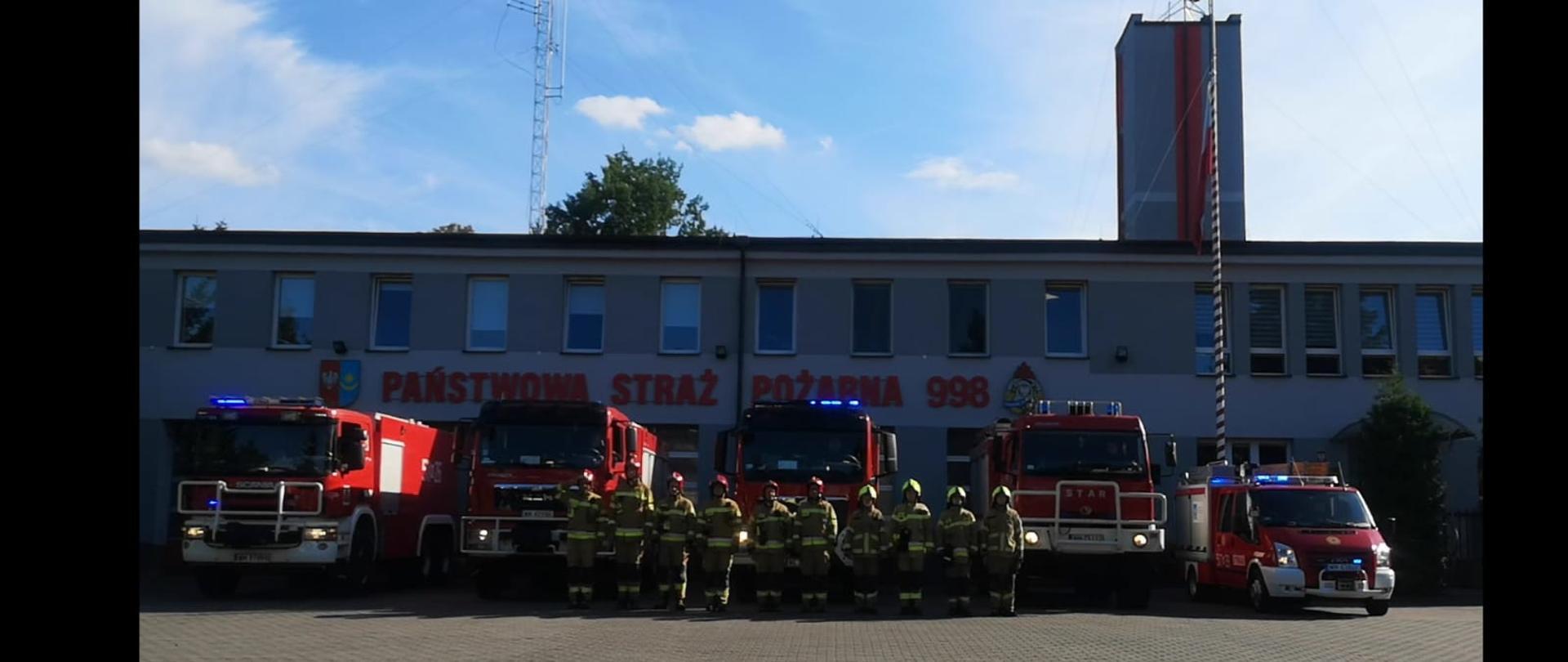 Funkcjonariusze KP PSP Mińsk Mazowiecki oraz ratownicy OSP Mińsk Mazowiecki na placu przed Komendą, za nimi samochody bojowe z włączonymi sygnałami świetlnymi oraz dźwiękowymi