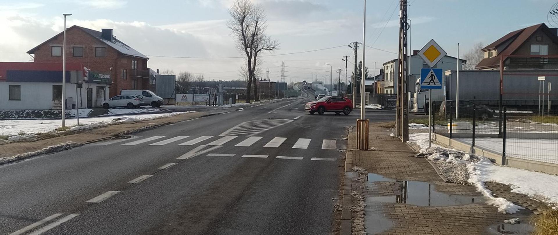 Droga przez miejscowość, zimowa aura. Na pierwszym planie widoczne przejście dla pieszych.