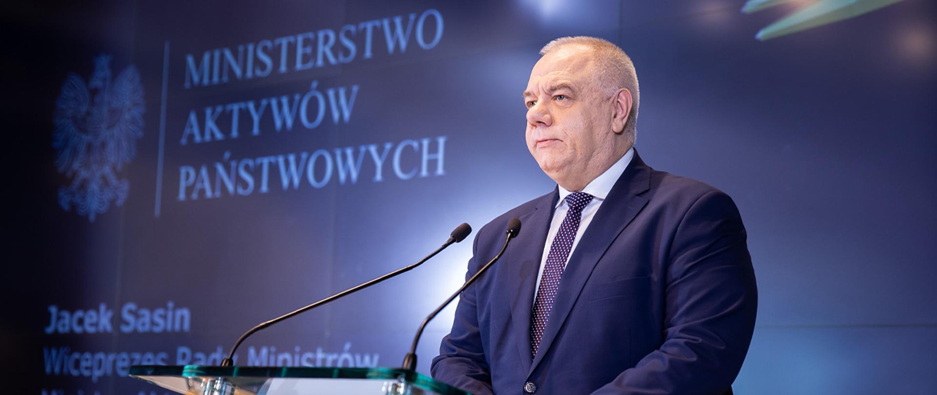 Wicepremier Jacek Sasin stoi przy mównicy. W tle ścianka z logotypem MAP oraz napisem Jacek Sasin, Wiceprezes Rady Ministrów, Minister Aktywów Państwowych