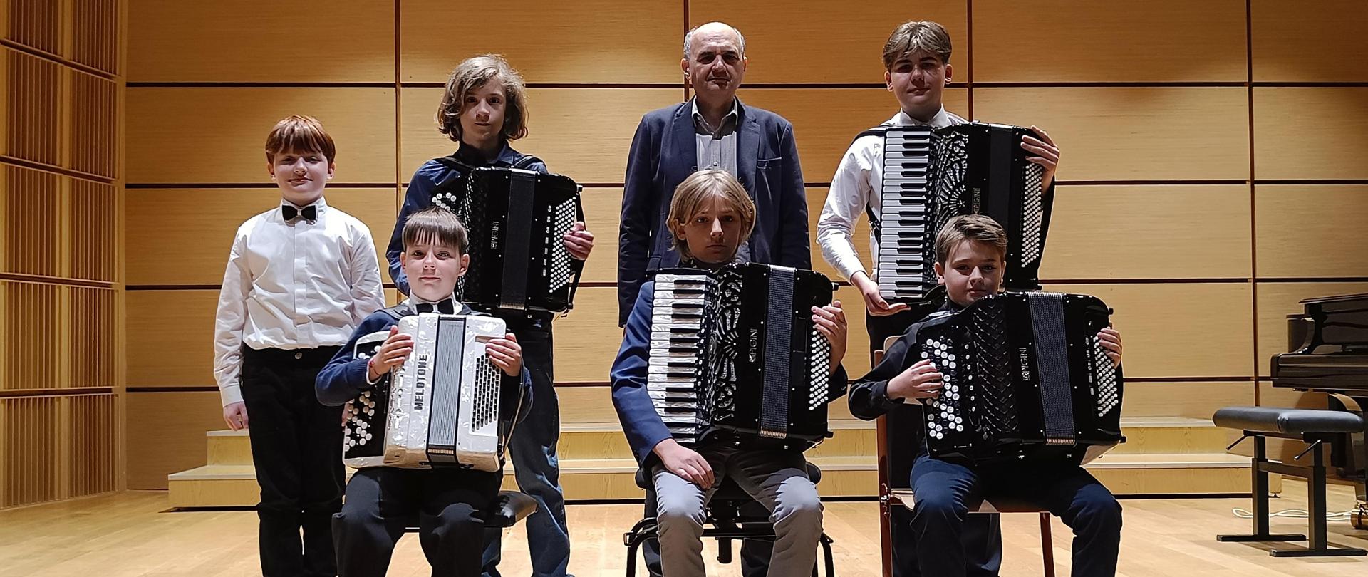 Z przodu trzech uczniów siedzących na krzesłach i trzymających akordeony. Z tyłu również trzech uczniów- dwóch z nich trzyma akordeony. Po środku stoi nauczyciel.